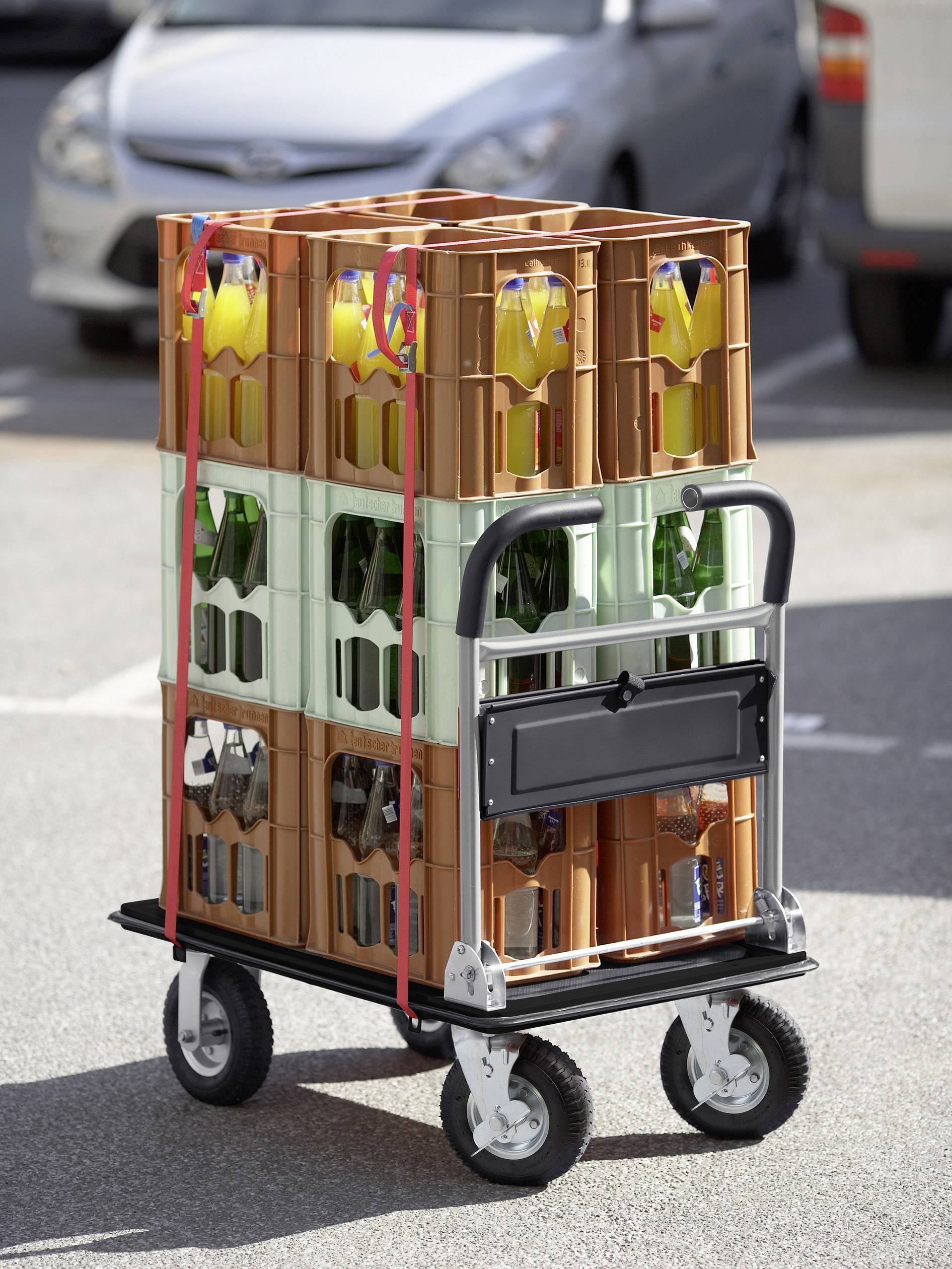 Flera dryckeslådor med flaskor på en handkärra, parkerad utomhus, troligen på en parkeringsplats.