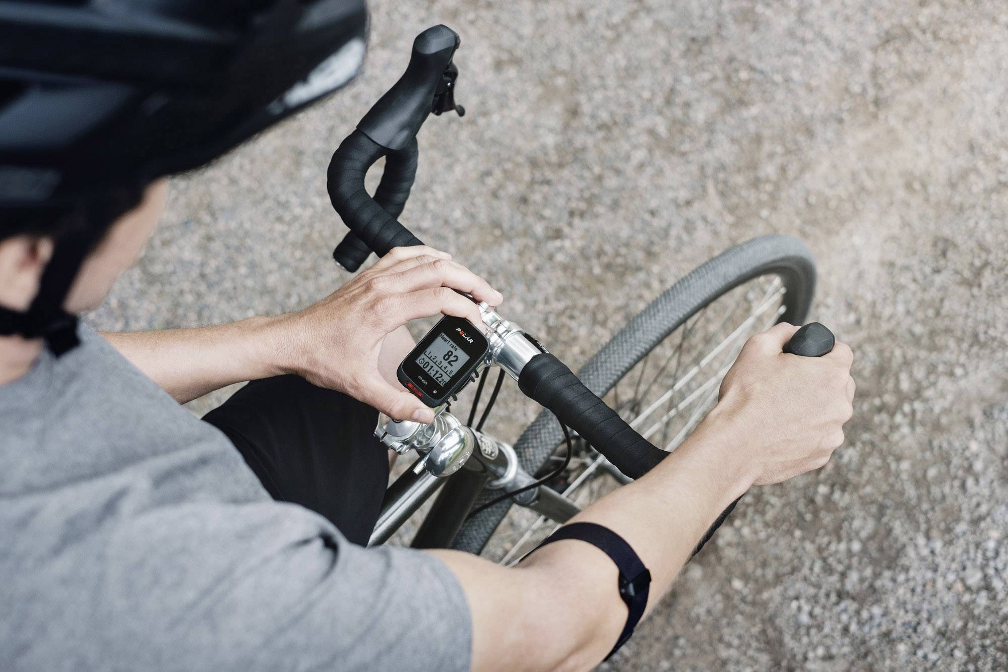 En cyklist i en grå tröja och hjälm som tittar ner på sin cykelkomputern. Displayen visar hastighet och kilometer. I bakgrunden syns en grusväg.