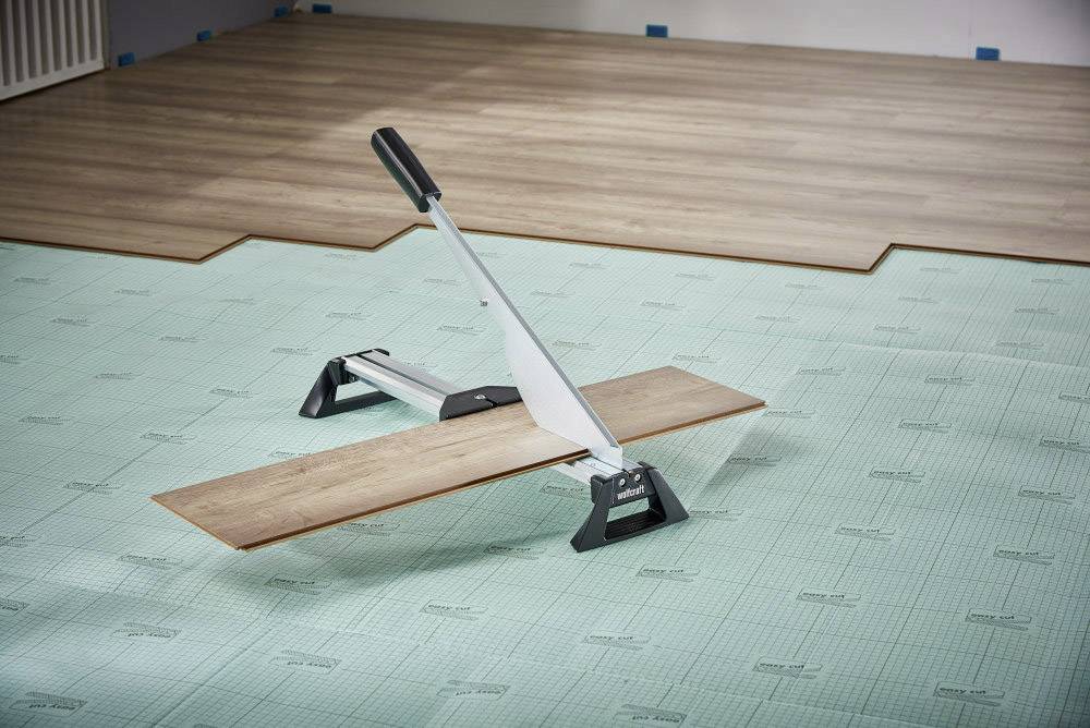 En laminatskärare på ett grönt underlag klipper en trälist; bakgrunden visar delvis installerade laminatpaneler.
