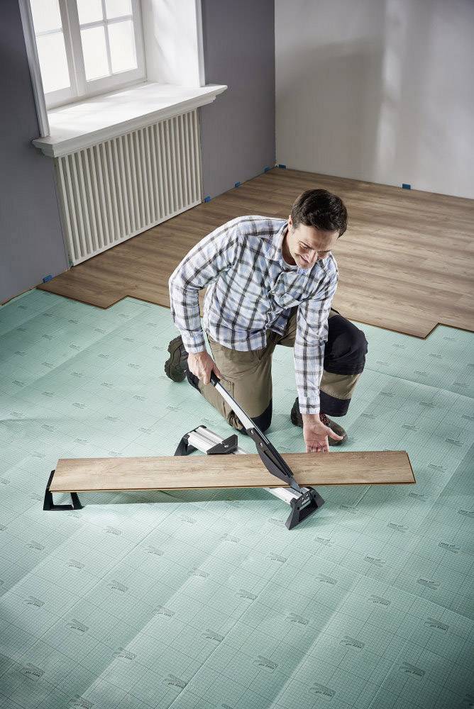 En person lägger laminatgolv i ett rum, knäböjer på ett skumunderlag och fäster en bräda med ett verktyg.