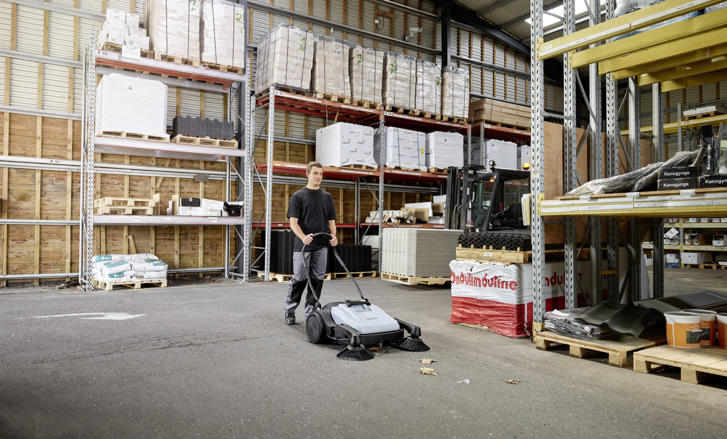 En man kör en industriell sopmaskin genom ett lager med höga hyllor, där lådor och material är placerade.
