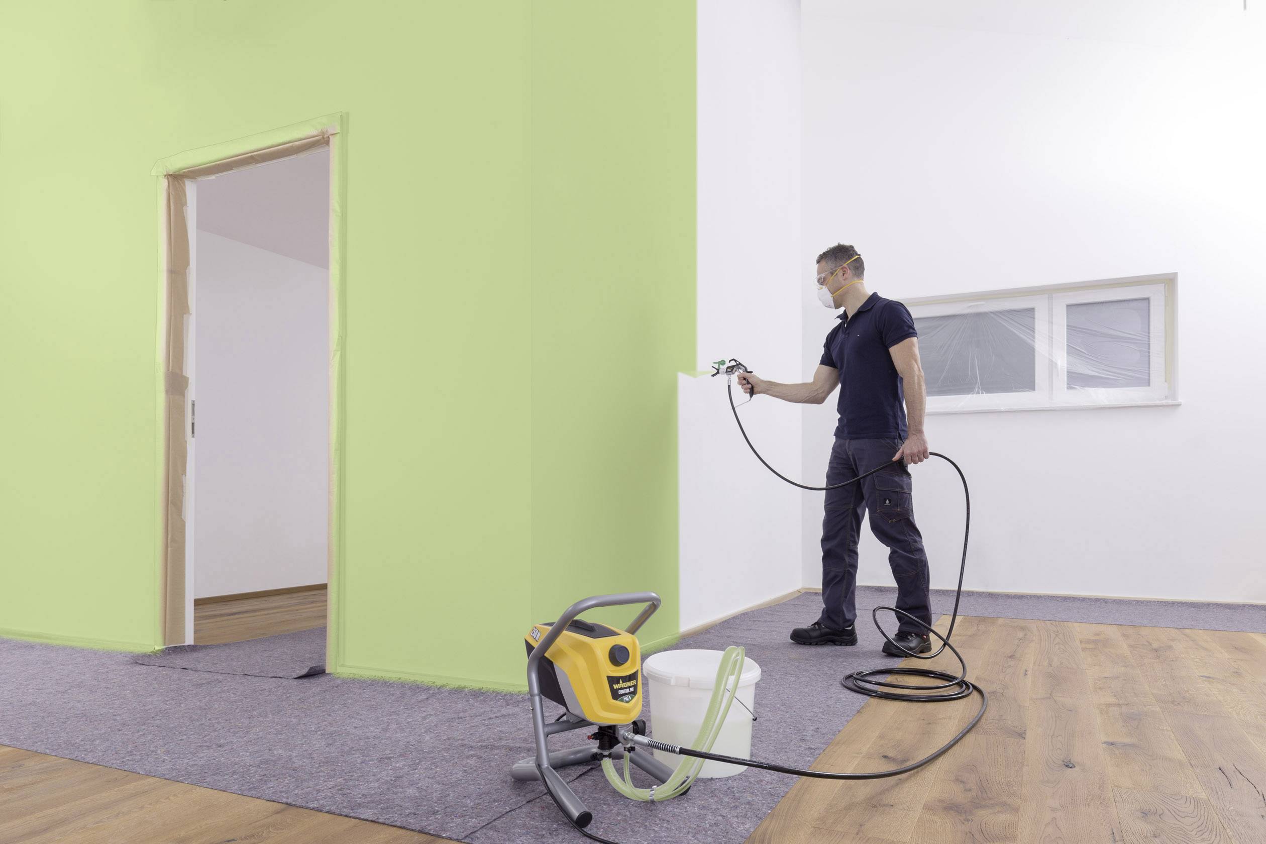 En man sprutar grön färg på en innervägg med en färgspruta. På golvet står en färgmaskin och en hink.