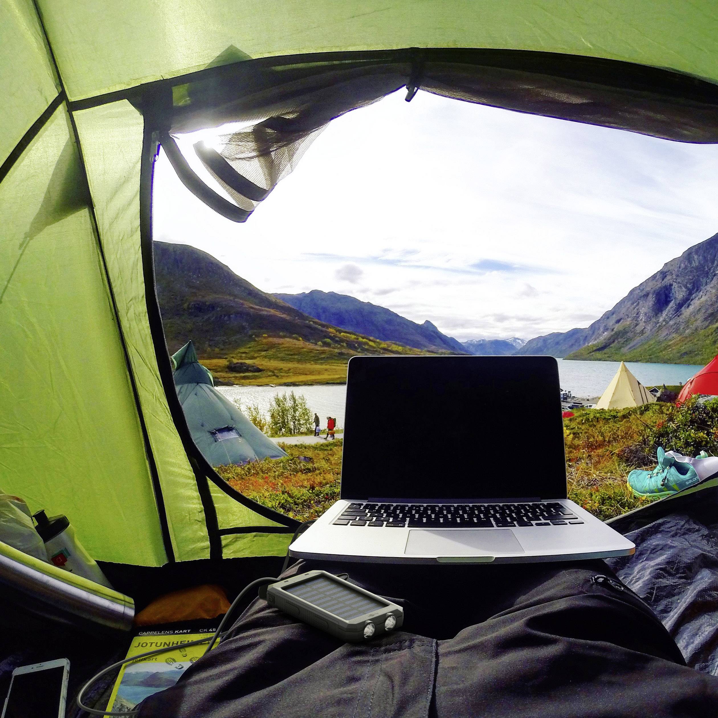 En person ligger i ett tält med en laptop på skötet och blickar ut över en pittoresk berglandskap med en sjö i bakgrunden.