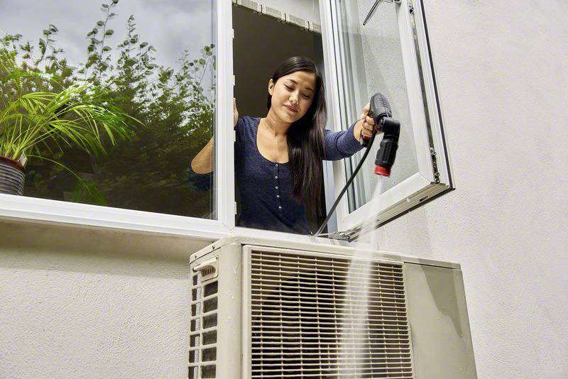 En kvinna rengör en utvändigt installerad luftkonditionering inifrån med en ångtvättare genom ett öppet fönster.