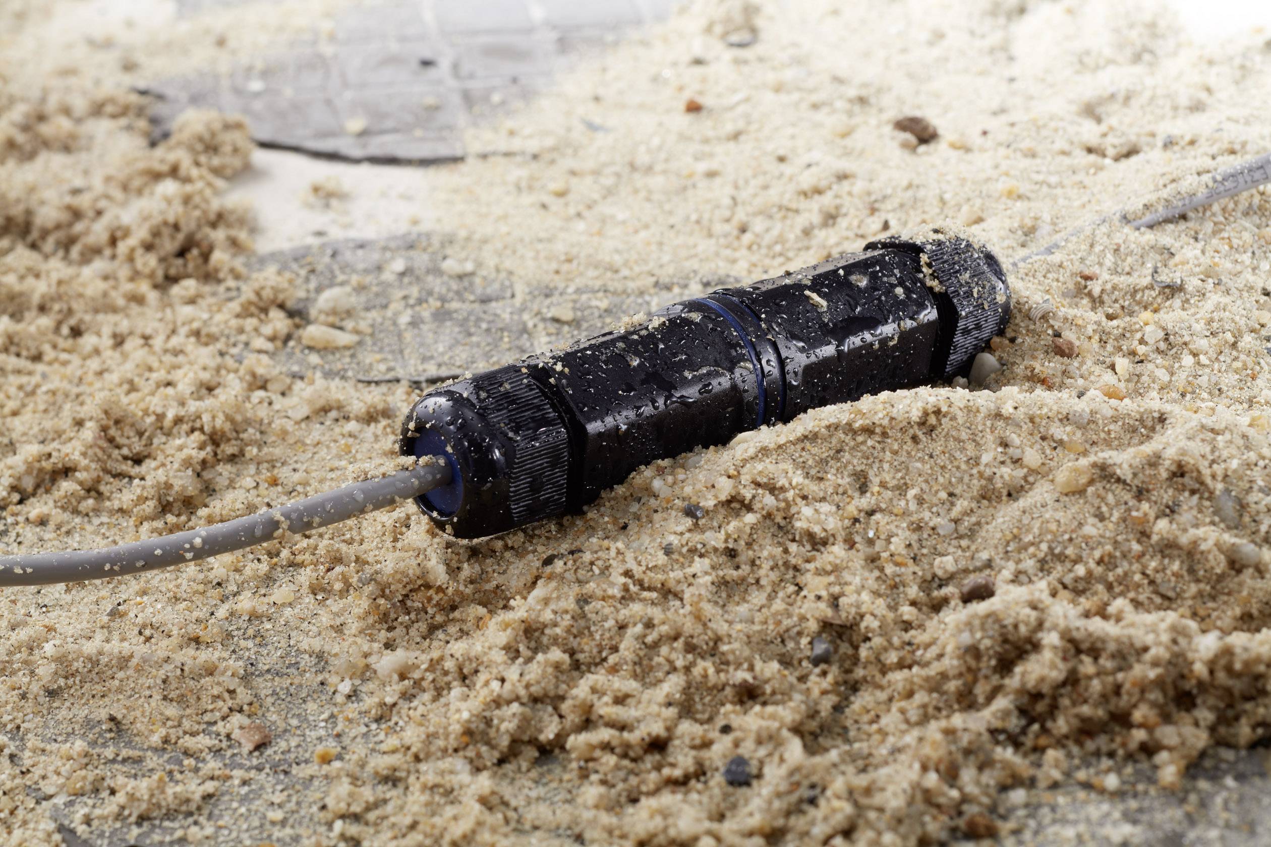 En svart kabel ligger på sandig mark, delvis täckt av sand, vilket tyder på en råare eller oskyddad miljö.