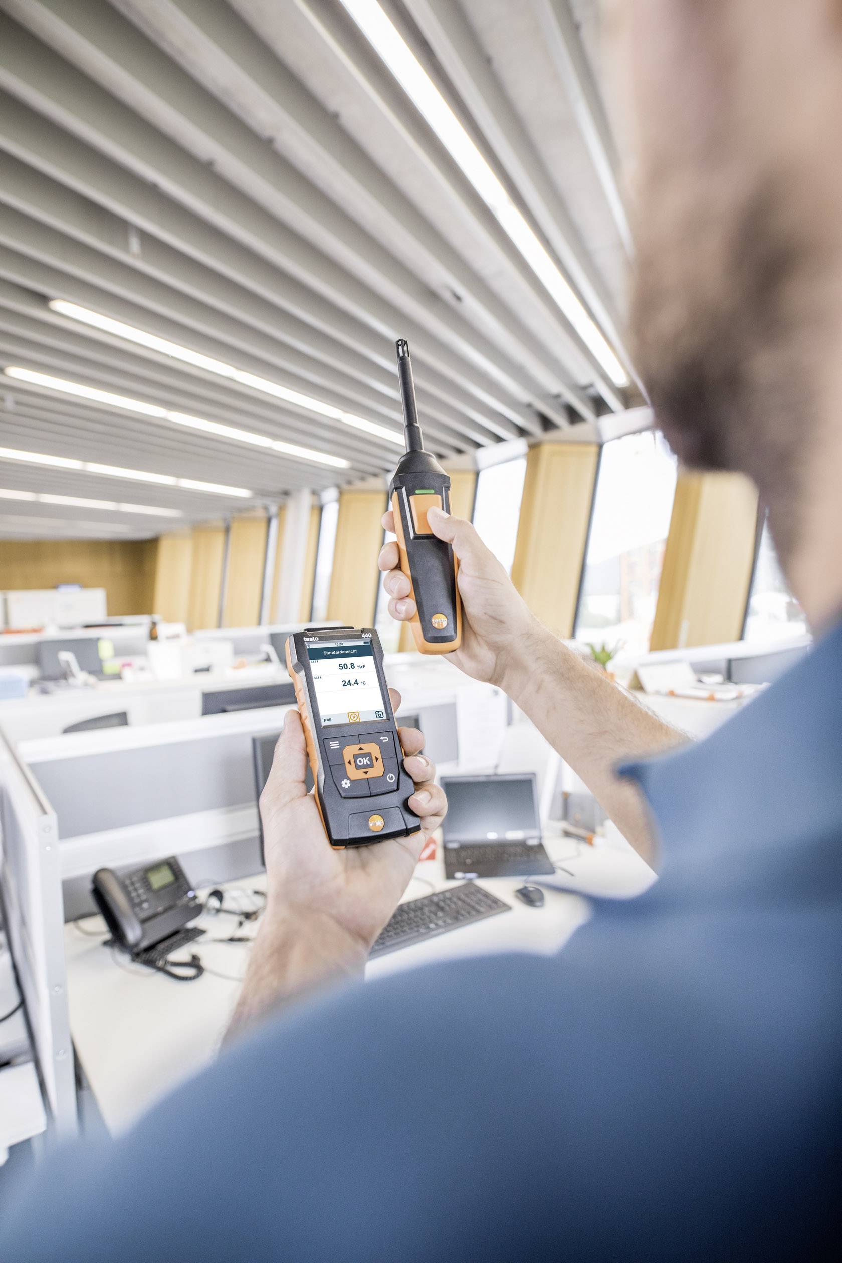 Man på kontoret använder ett mätinstrument för att kontrollera luftkvaliteten på en arbetsplats. Flera skrivbord och datorer syns i bakgrunden.