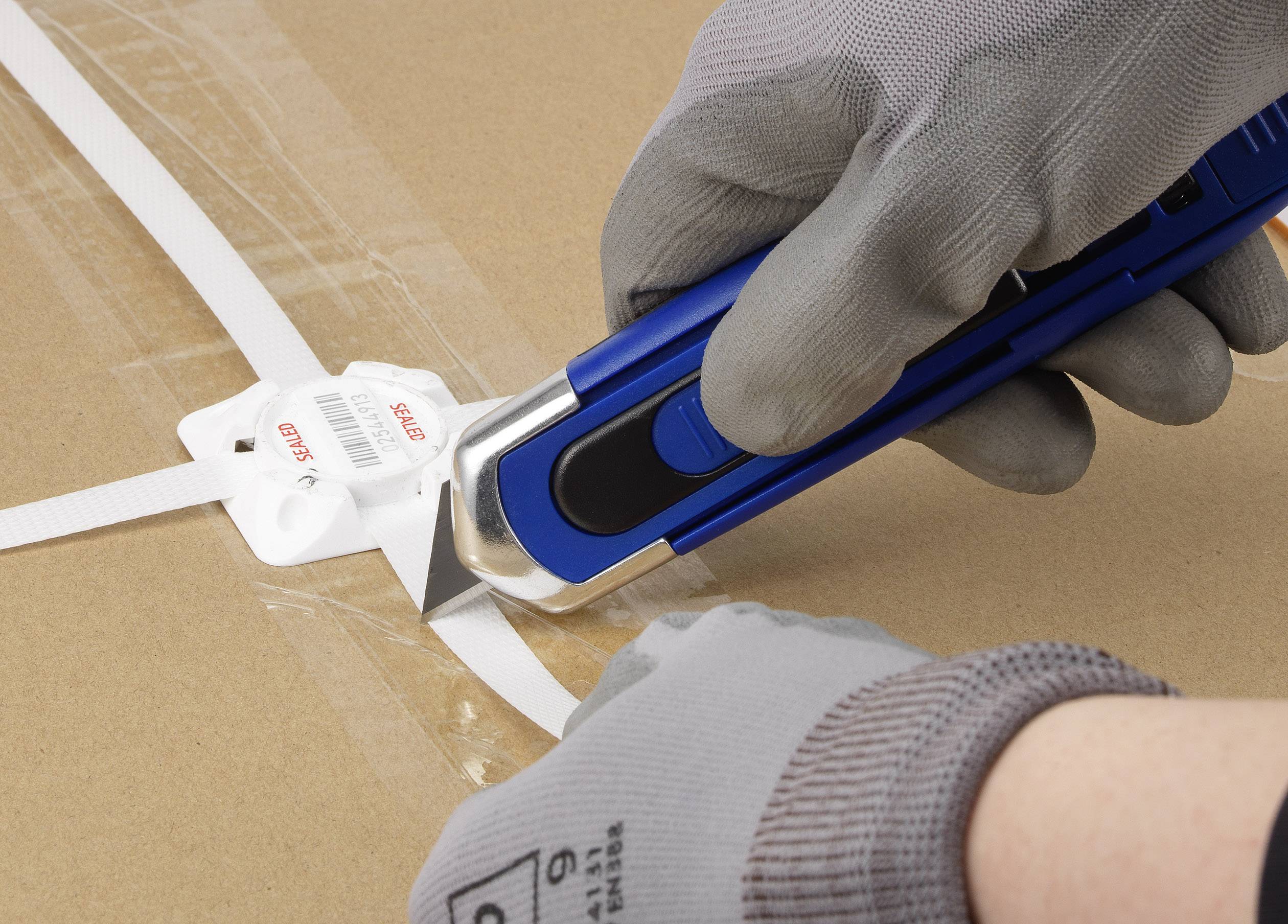 En person klipper ett vitt plastband på en kartong med en blå skärkniv. Personen bär grå skyddshandskar.