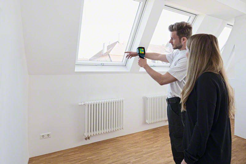 En person visar en värmekamera för någon annan i ett rum med takfönster och radiatorer i syfte att eventuellt identifiera värmläckage.