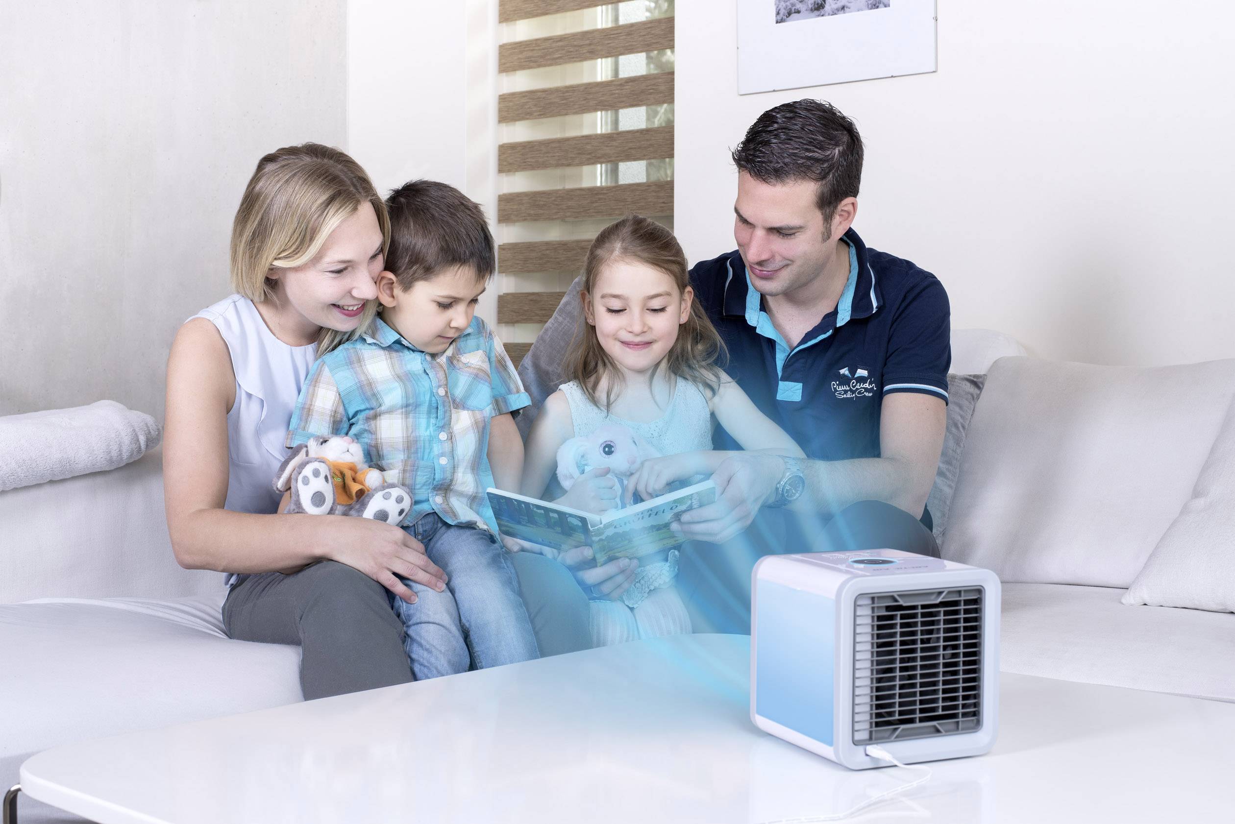 En familj sitter tillsammans på en soffa och läser en bok. Framför dem står en liten bordsbordsventilator som blåser sval luft.