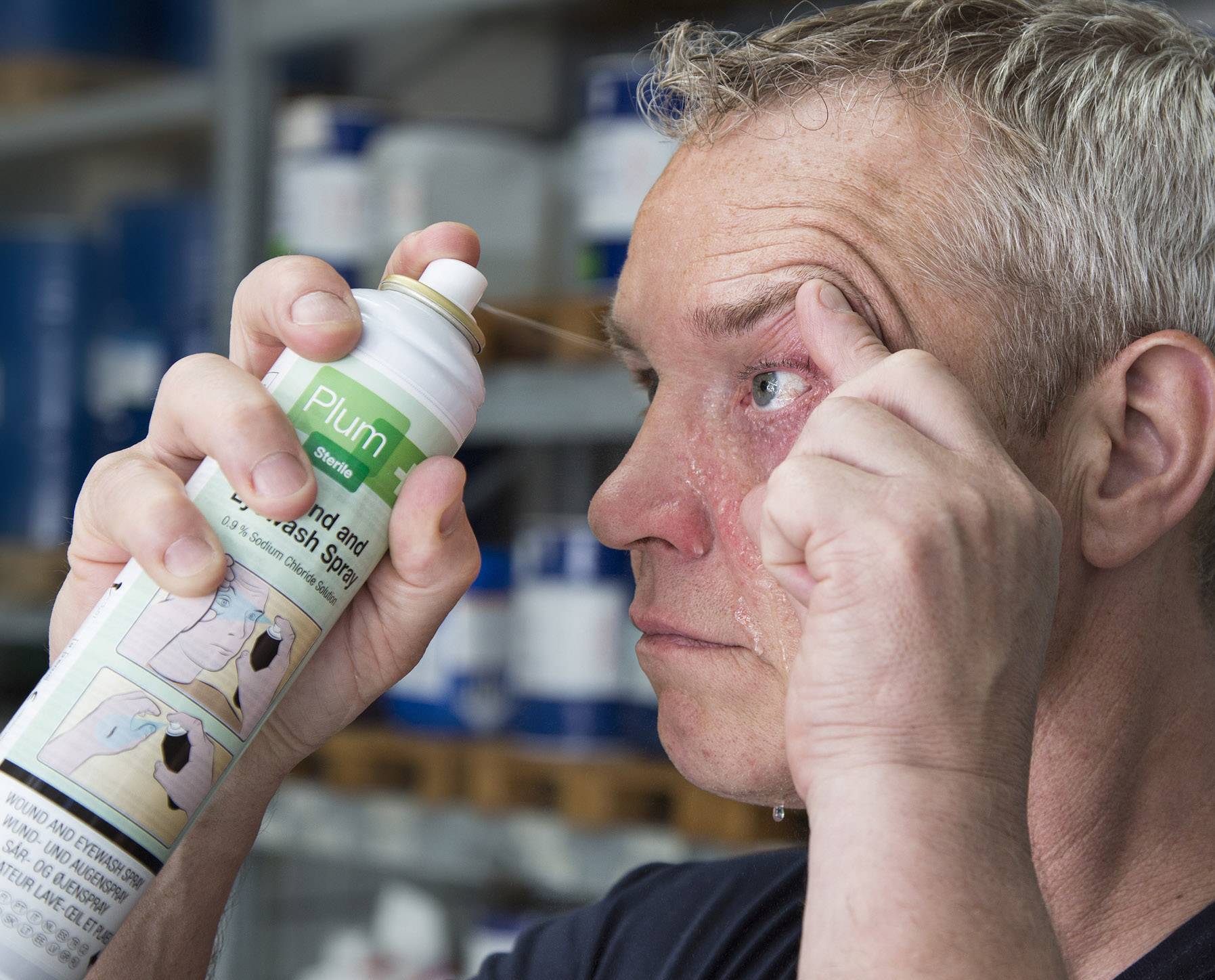 En man sprutar ögontvättsvätska i sitt öga för rengöring. Vätskan droppar från hans ansikte. I bakgrunden finns hyllor med produkter.