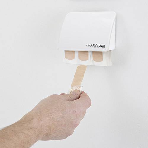 En hand tar ut ett plåster från en vit plåsterdispenser på väggen. Dispensern visar tre synliga plåster.