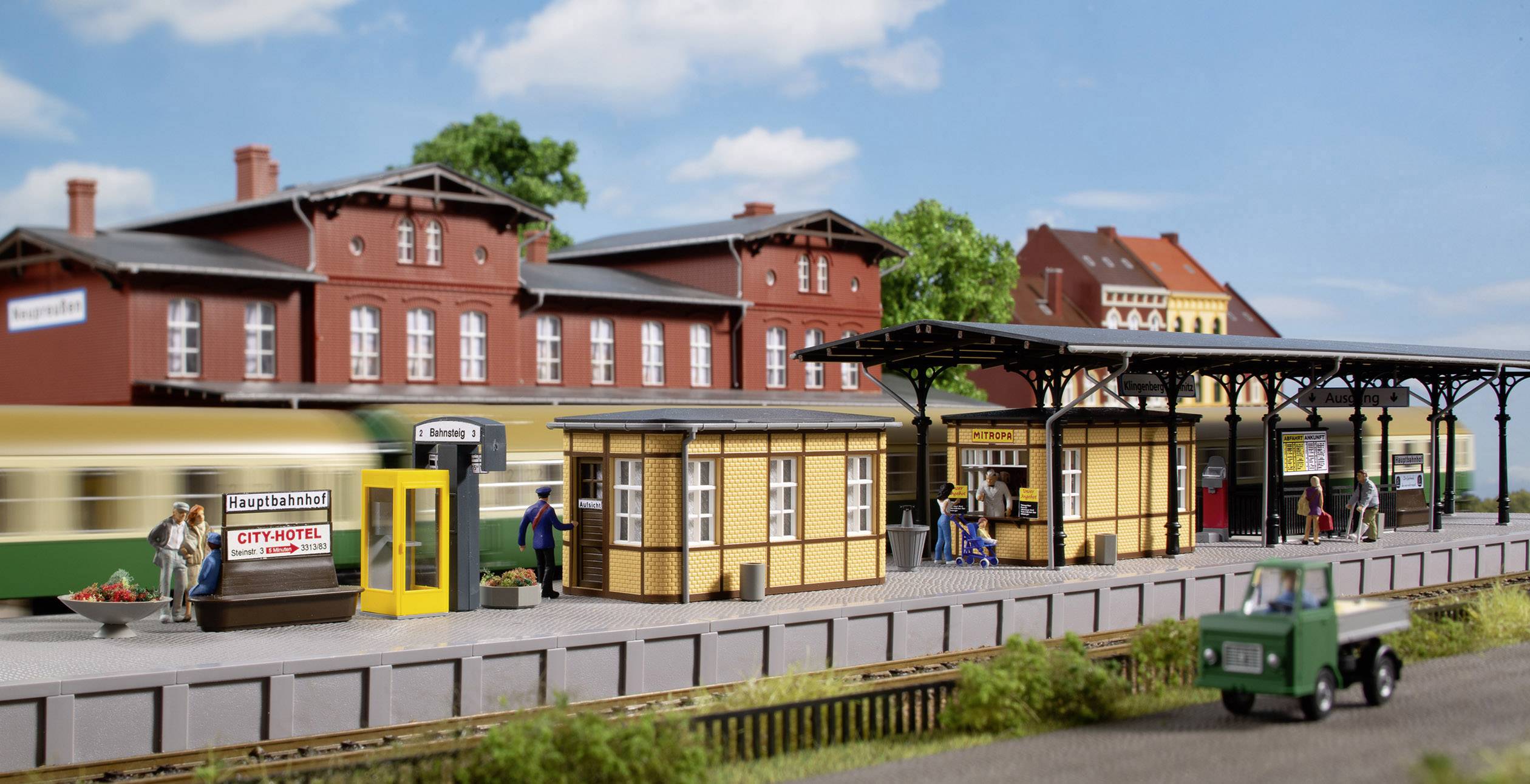 En modelljärnvägsstation med en historisk byggnad, flera figurer och ett gammalt tåg. Ett grönt landskap i bakgrunden.