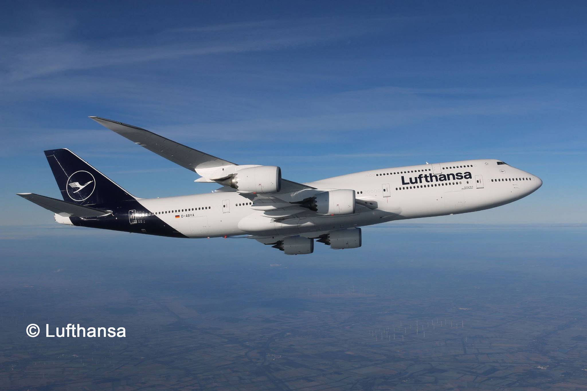 Ett stort flygplan från flygbolaget Lufthansa flyger på hög höjd över ett landskap, synligt mot en klarblå himmel.