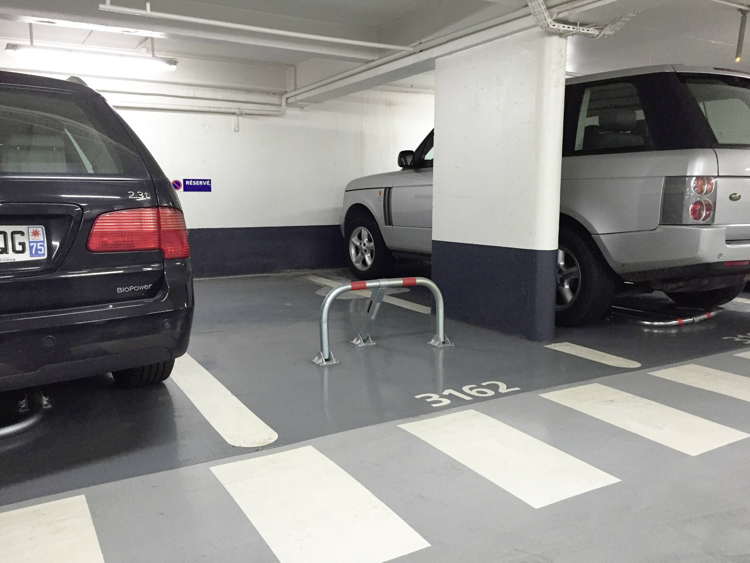 Två parkerade bilar i ett parkeringsgarage. En parkeringsplats emellan är spärrad med en bom. Markeringen på golvet visar '3162'.
