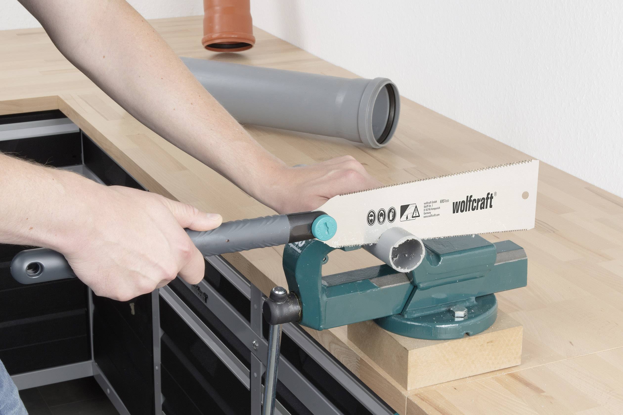 En person sågar ett grått plastror i en verkstad. Röret är fastspänt i ett skruvstäd.