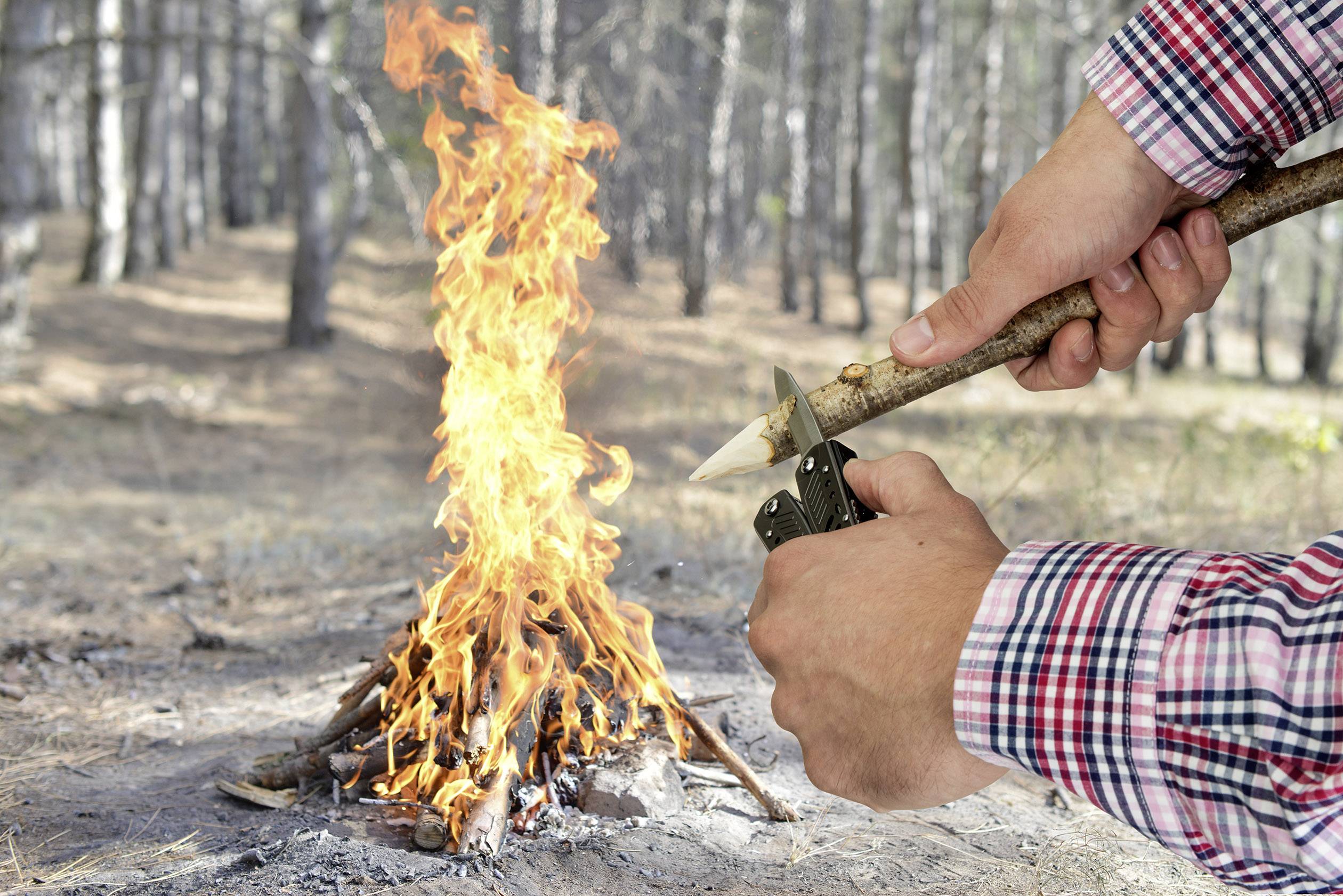 En person täljde med en kniv en gren i en skog, medan en lägereld brann i bakgrunden.