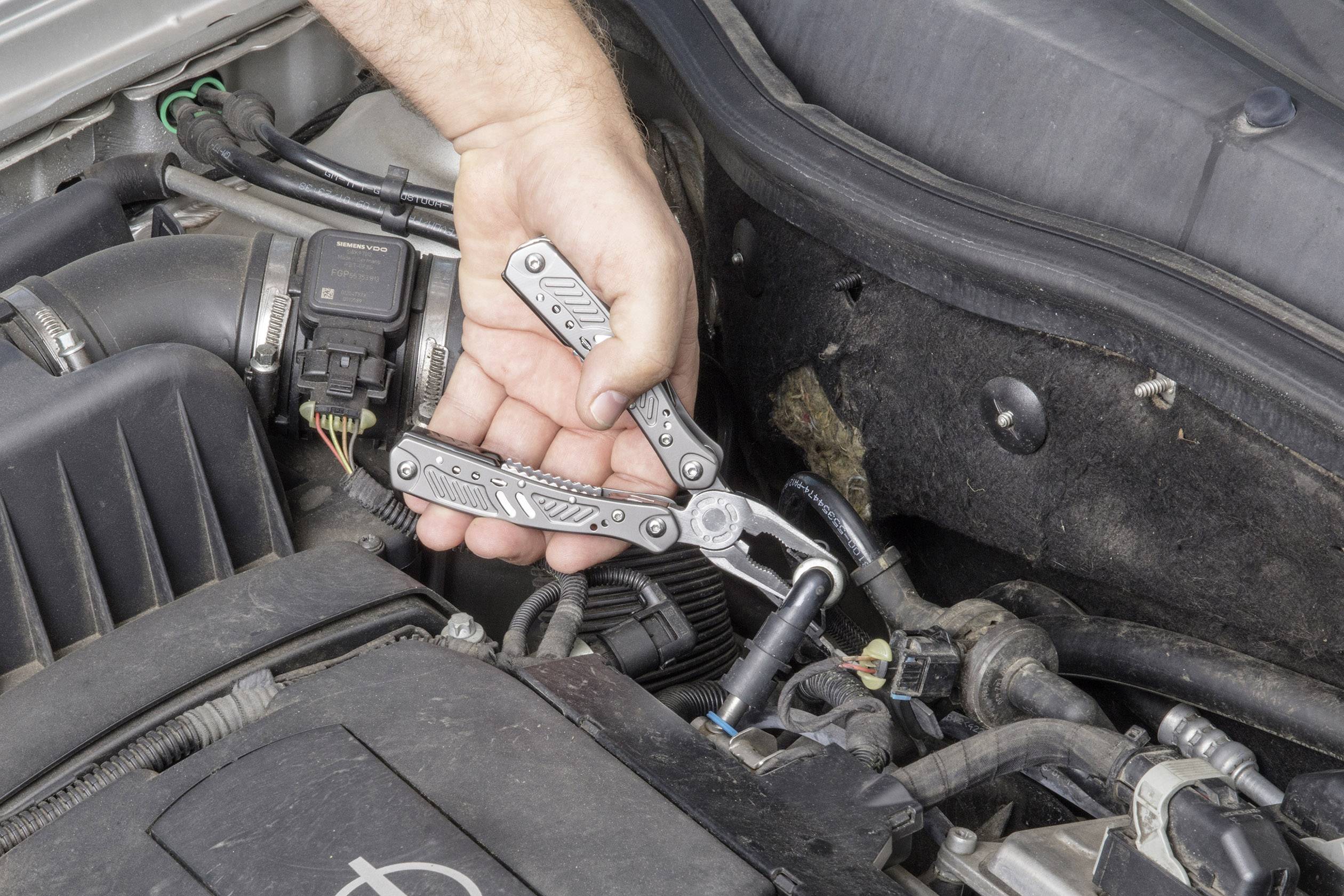 En person använder ett multiverktyg för att reparera en del av motorn på en bil. Bilen är öppen, och fokus ligger på verktyget.