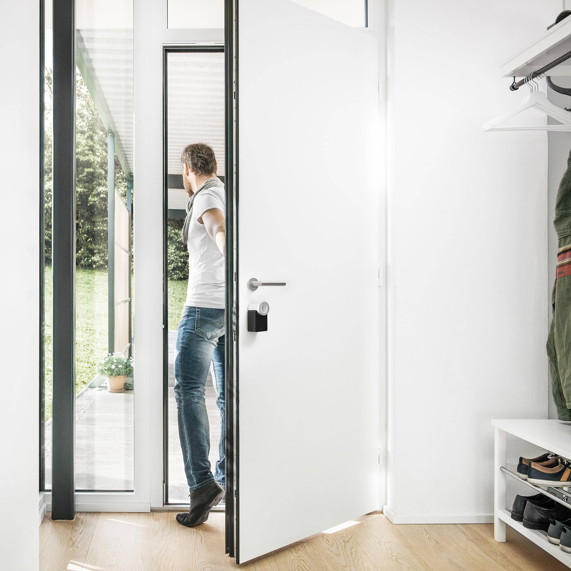 En man står i dörröppningen till ett modernt hus och blickar ut över en grön trädgård; ljus, minimalistisk inredning.