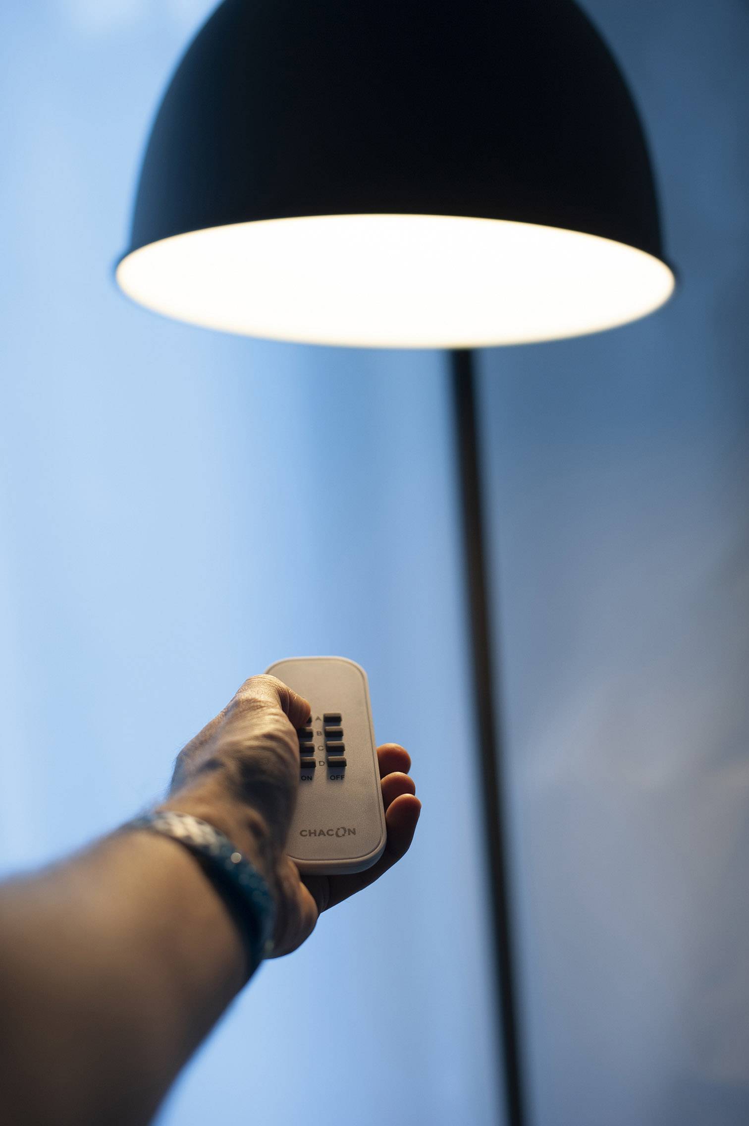 En hand håller en fjärrkontroll framför en tänd lampa. Lampan lyser upp rummet jämnt uppifrån och ned.