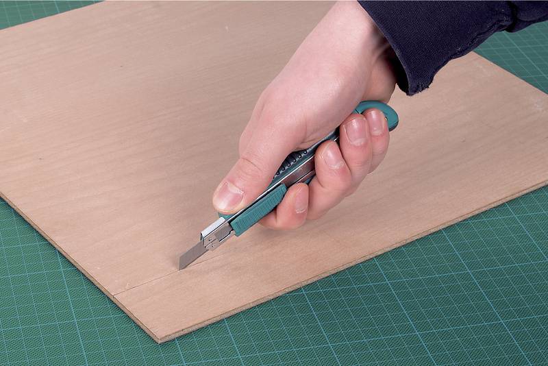 En hand skär en tunn träskiva med en cutterkniv på en grön skärmatta.