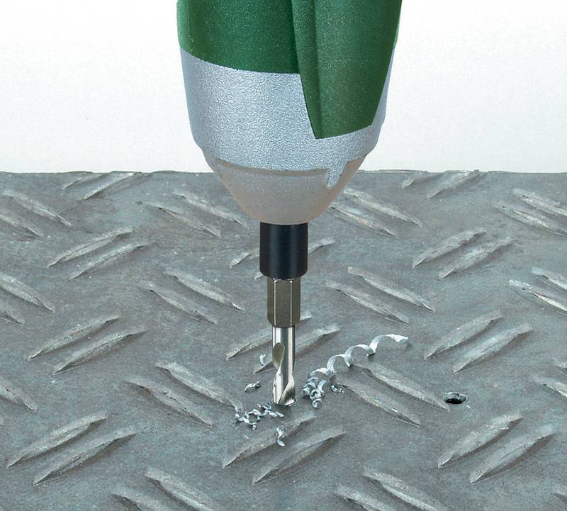 En grön elektrisk borr borrar ett hål i en metallplatta med diamantmönstring, omgiven av metallskärspån.