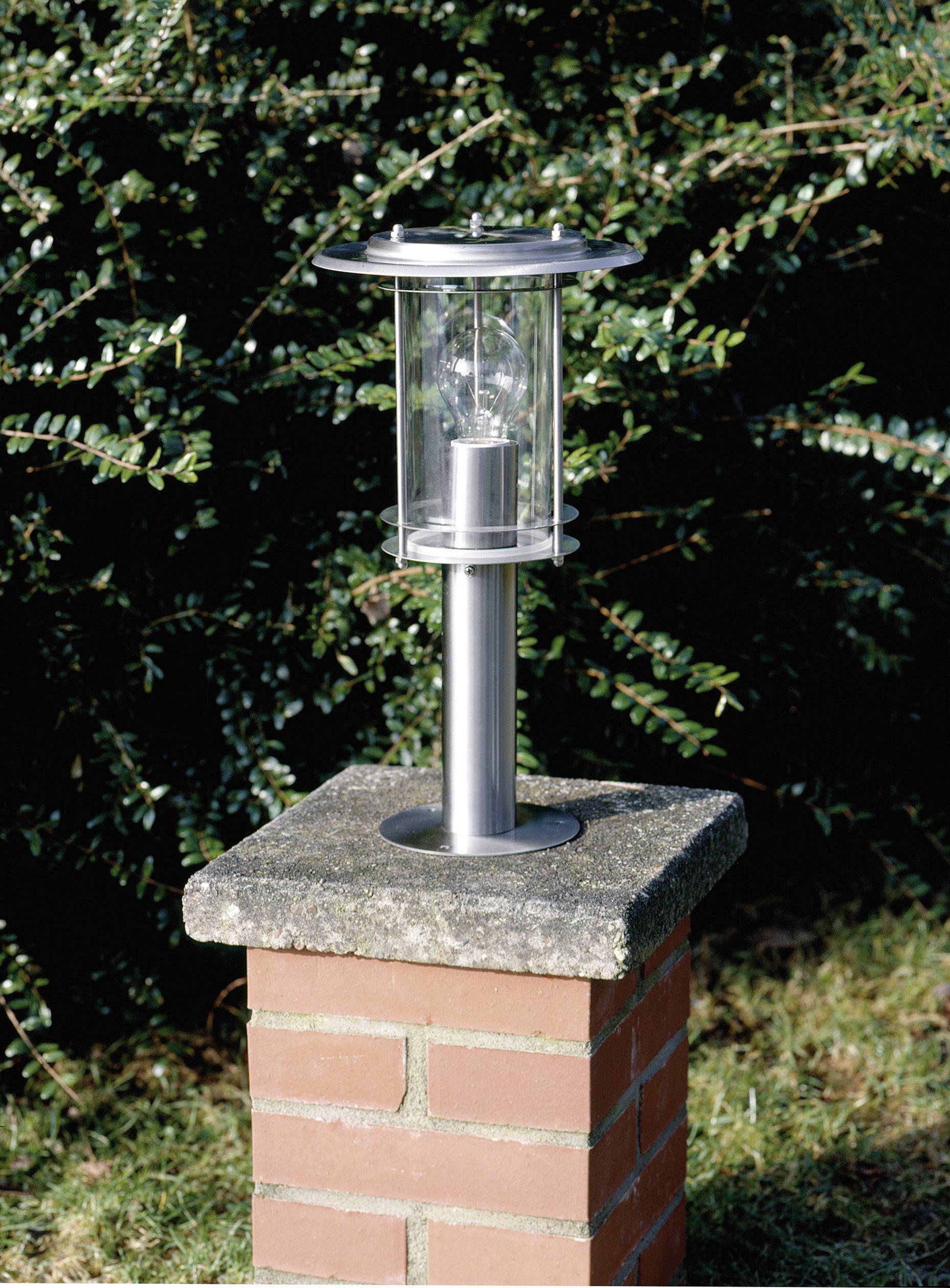 En silvergrå trädgårdslampa på ett murat fundament framför en bakgrund av gröna blad.