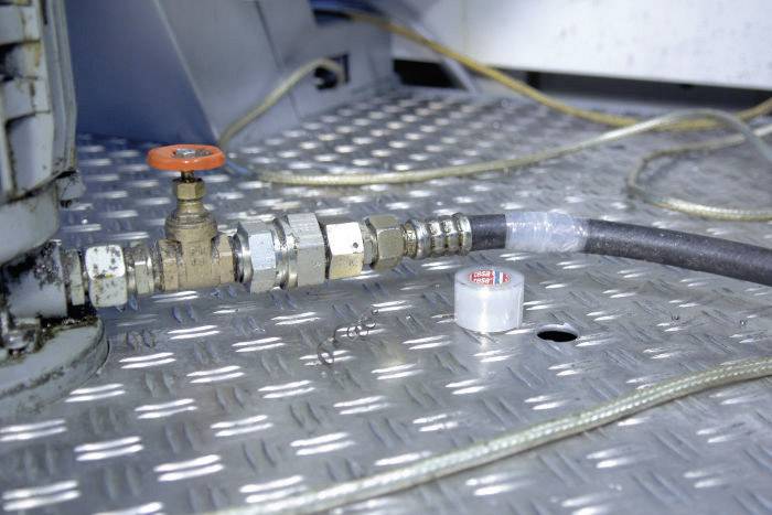 En vattenkran med ett rött handtag och en ansluten slang på en räfflad metallyta, med kablar som löper i bakgrunden.