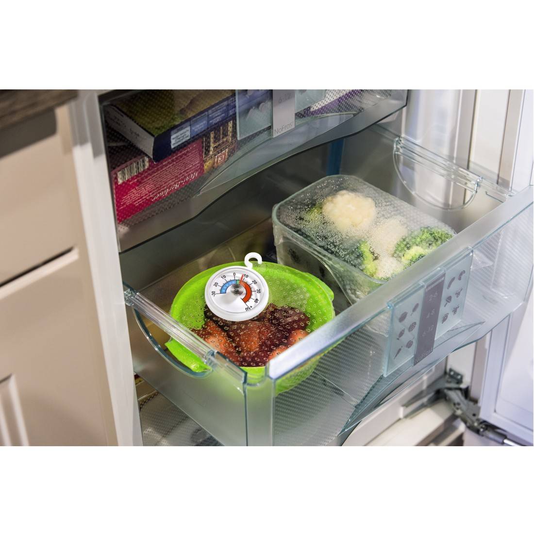 Ett kylskåpsfack med en skål röda bär och en termometer. Bredvid står en skål med broccoli och blomkål.