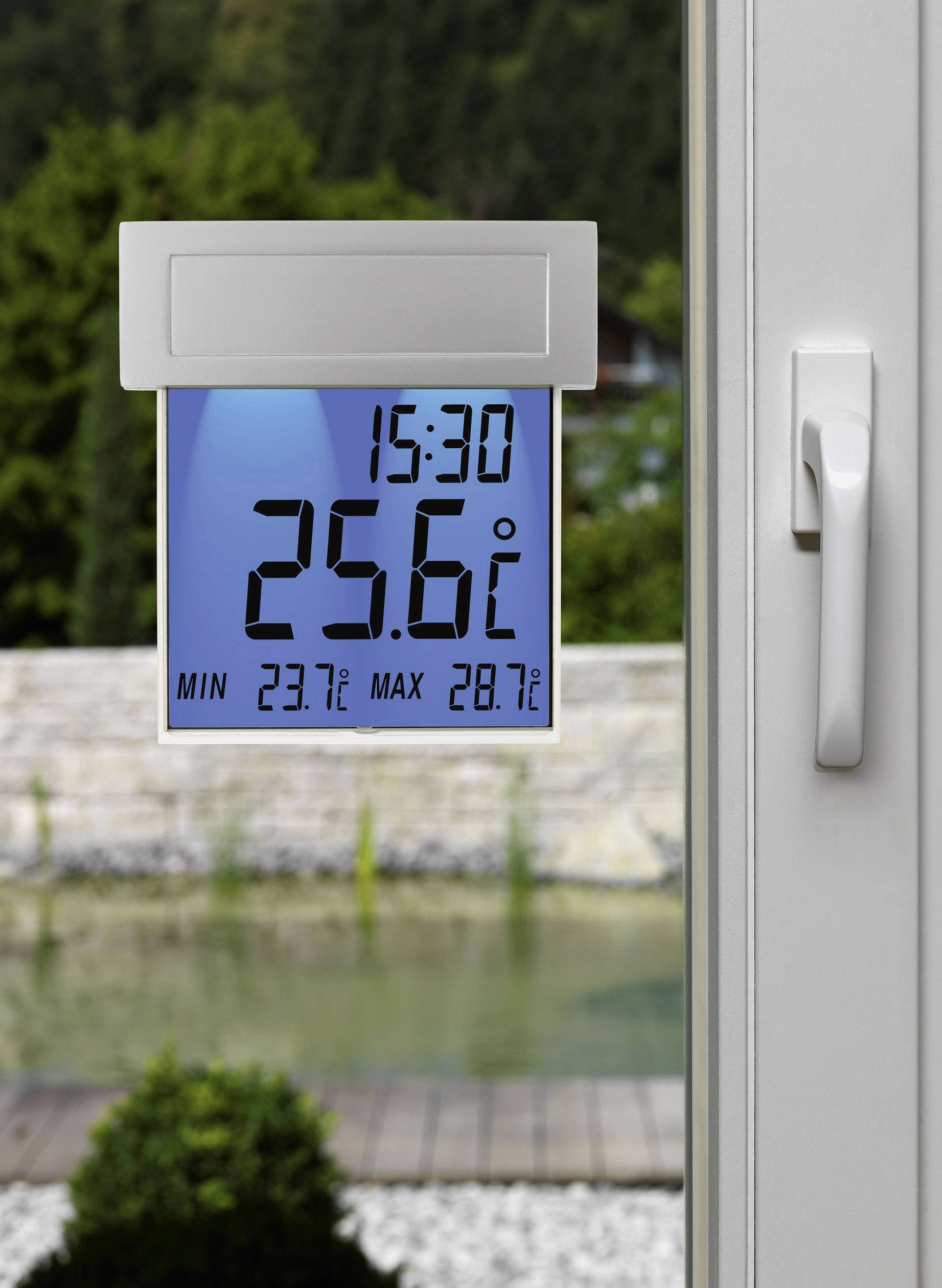 Digitalt termometer visar 25,6°C klockan 15:30. Min 23,7°C, Max 28,7°C. Bakgrund: Trädgård med damm, sedd genom ett fönster.