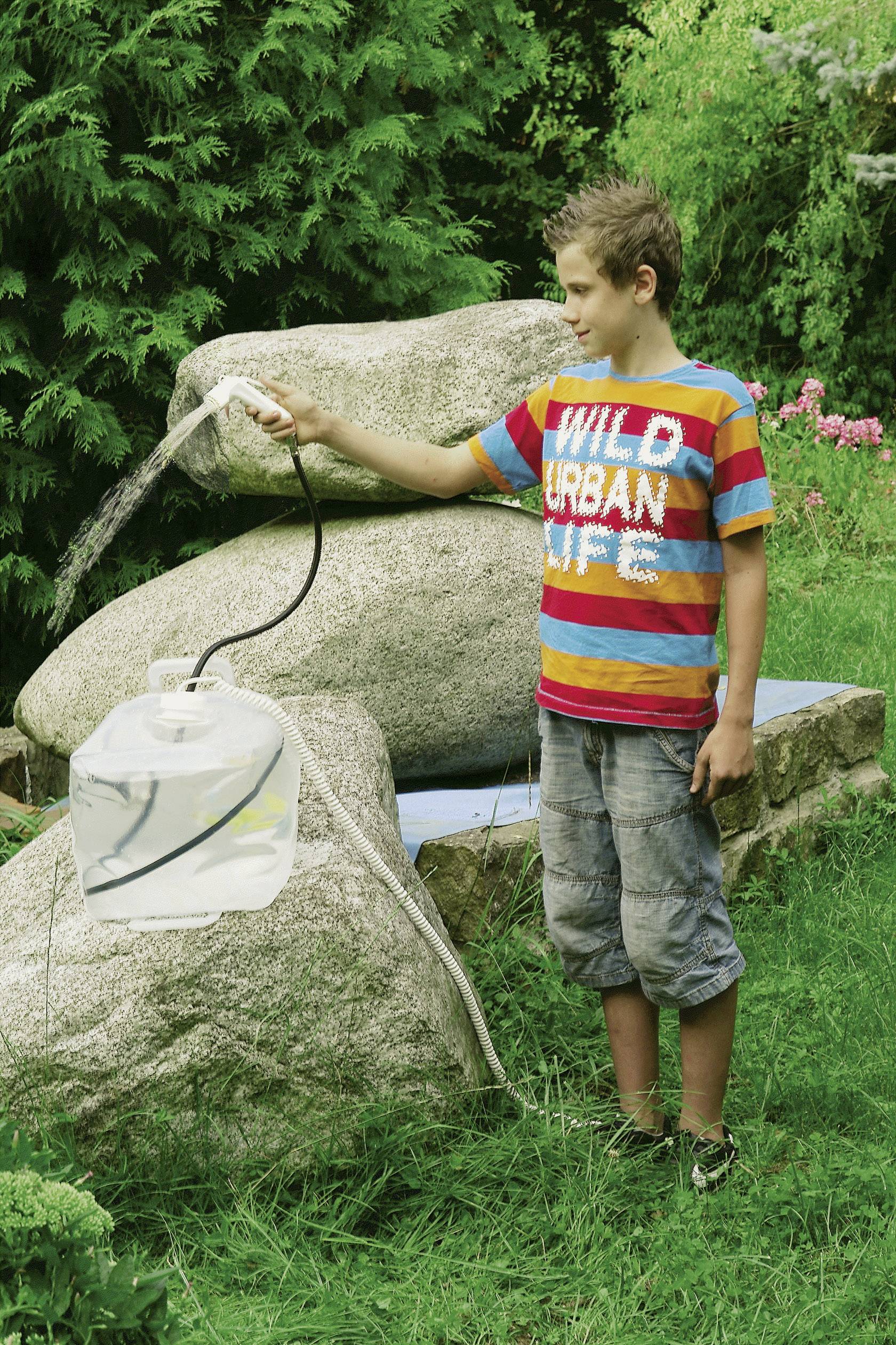 En pojke i en randig t-shirt häller vatten från en slang i en klar behållare. I bakgrunden finns gröna växter.
