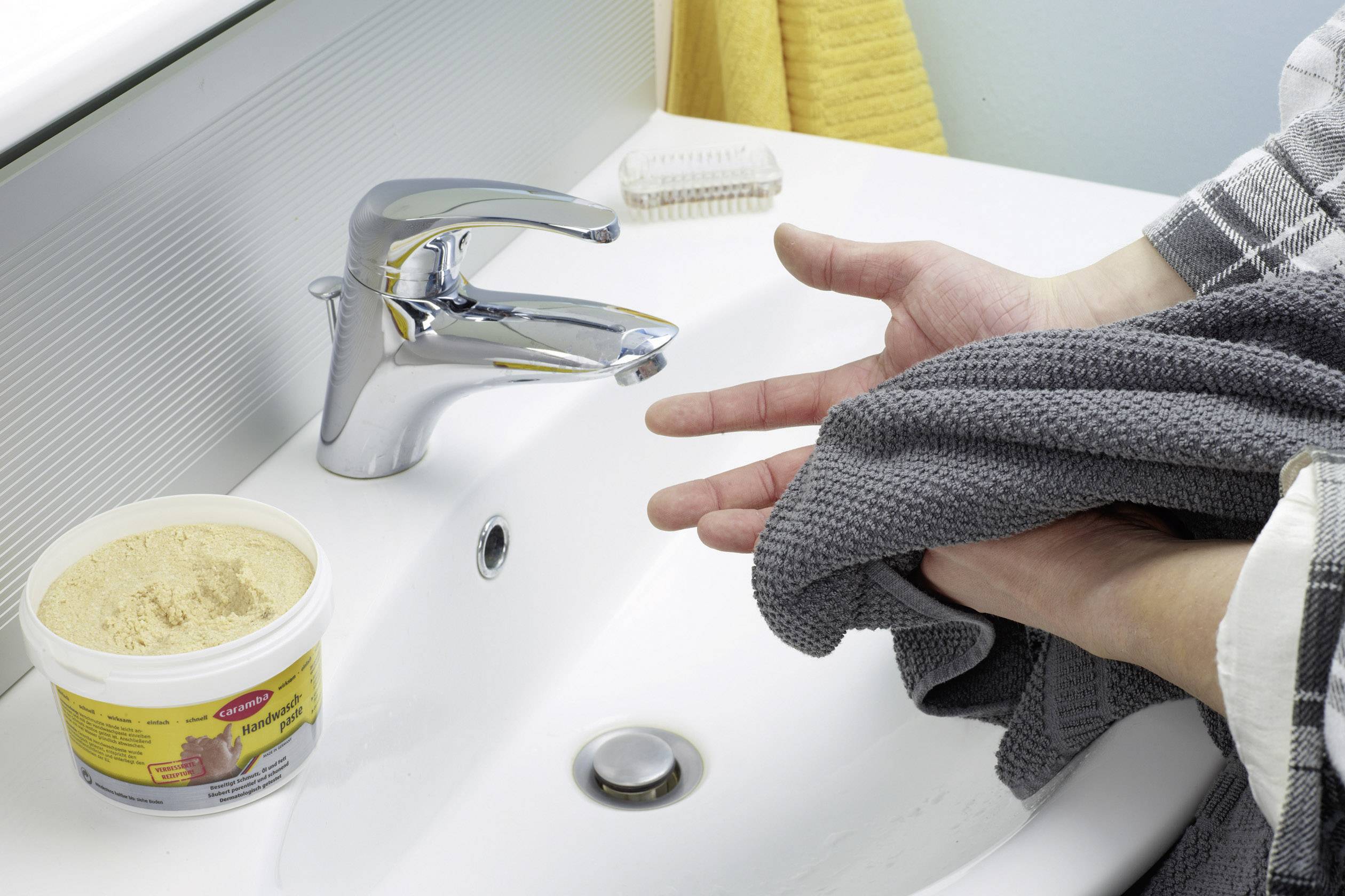 En person torkar händerna med en grå handduk intill ett handfat med en kran och en behållare med rengöringsmedel.