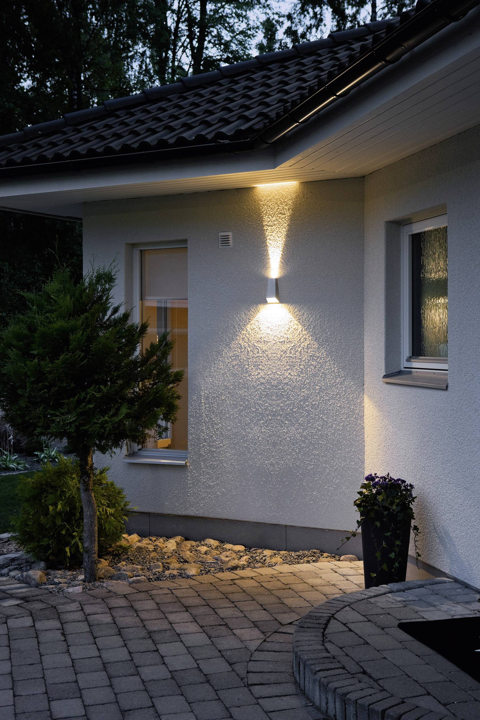 Två fönster på ett hus med utomhusbelysning på väggen, som lyser upp en stenlagd gång och en liten växt bredvid fasaden.