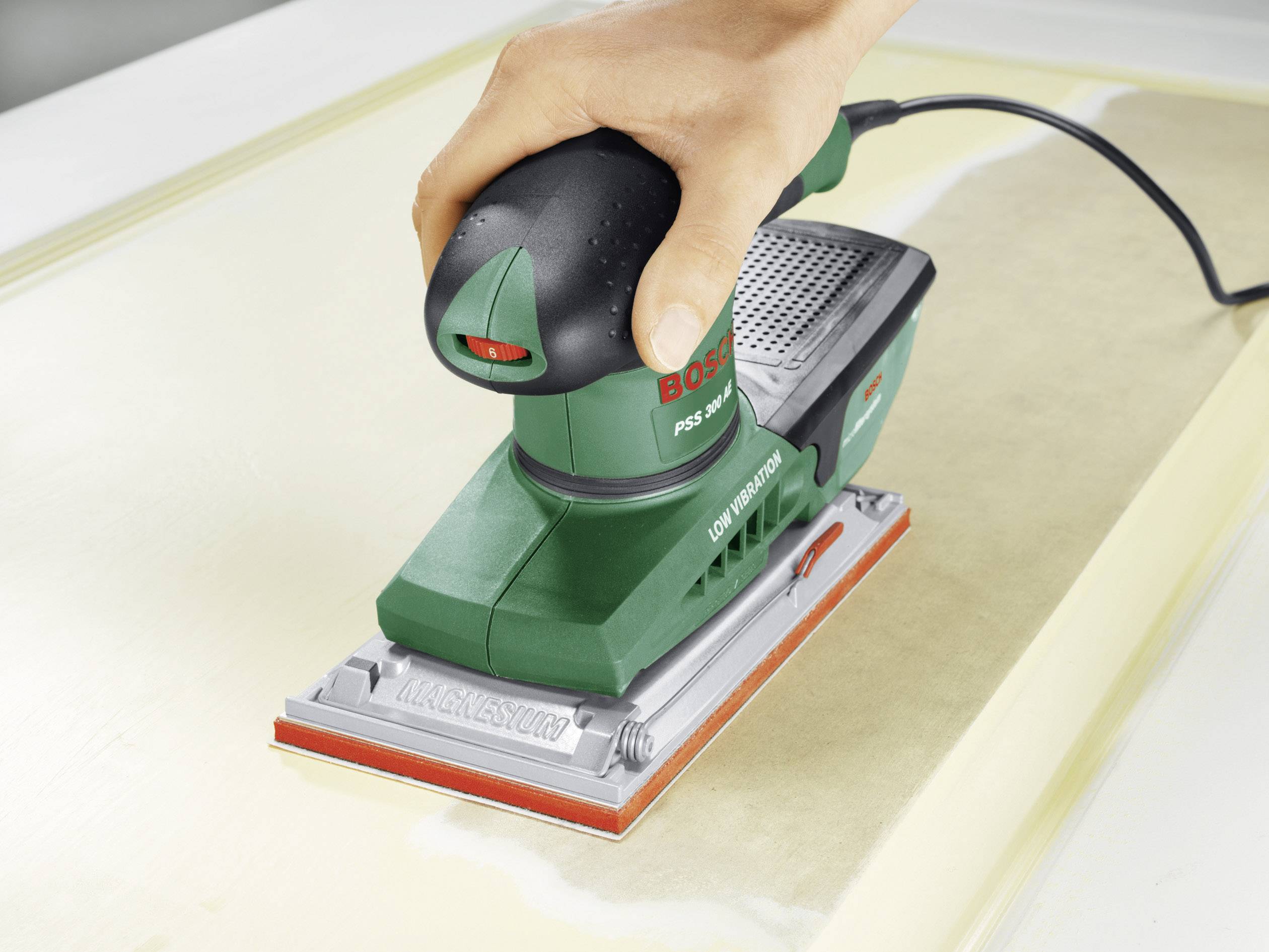 En hand håller en grön Bosch-slipmaskin som arbetar på en träyta. Slipmaskinen är elektrisk och ansluten med en sladd.