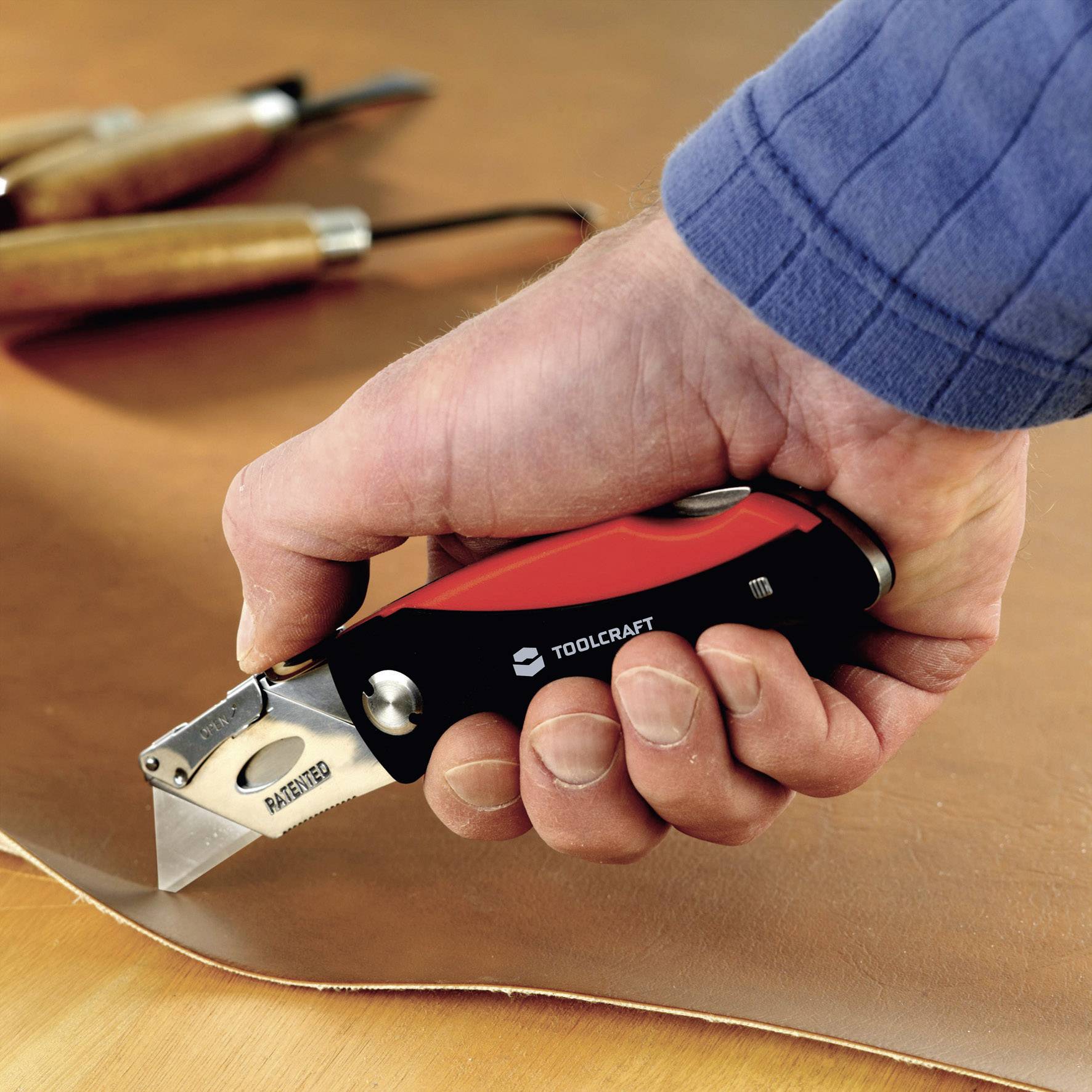 En hand håller en vass mattkniv och skär ett stycke tjockt läder. I bakgrunden syns träverktyg.