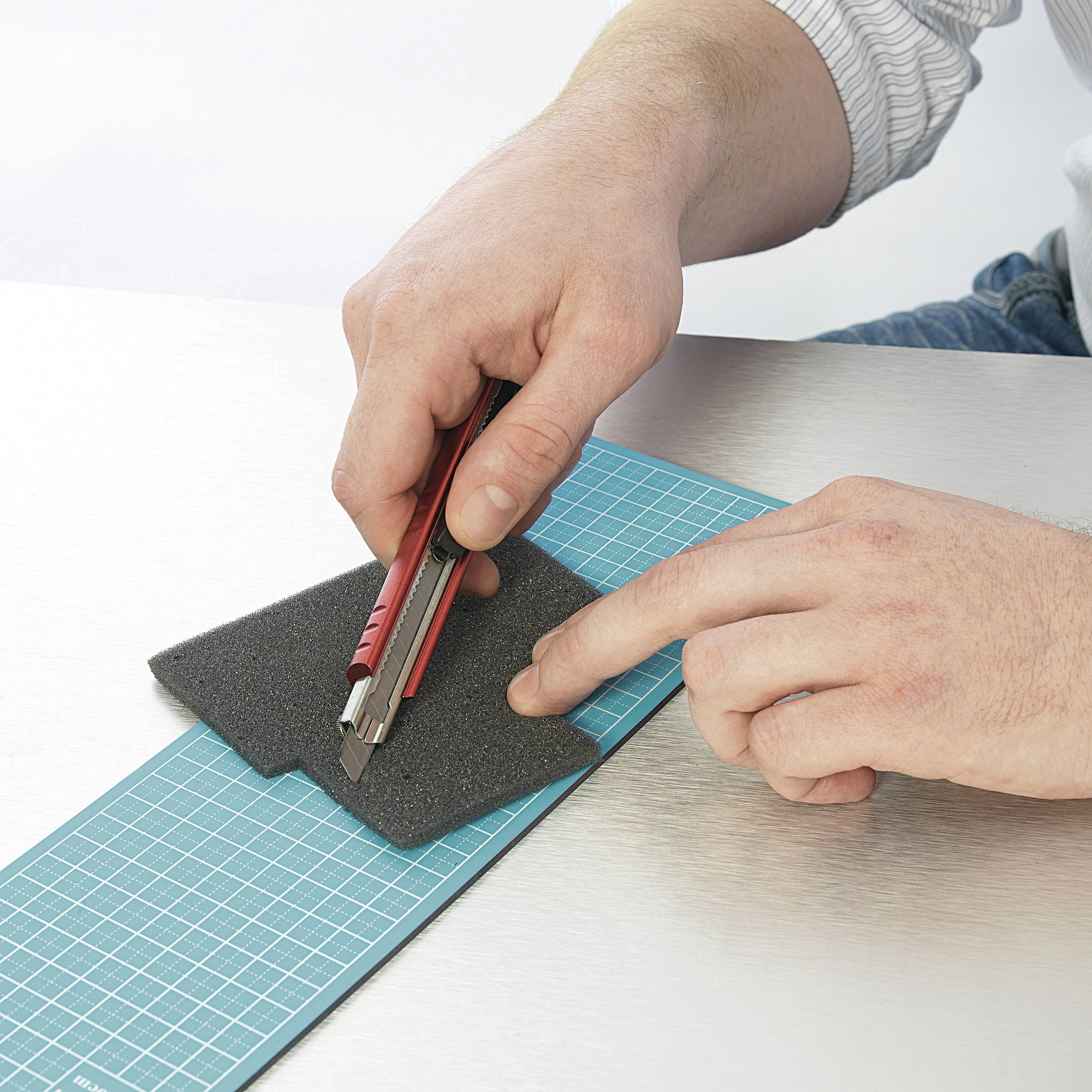 En hand skär ett svart skumstycke med en röd cutterkniv på en skärmatta.