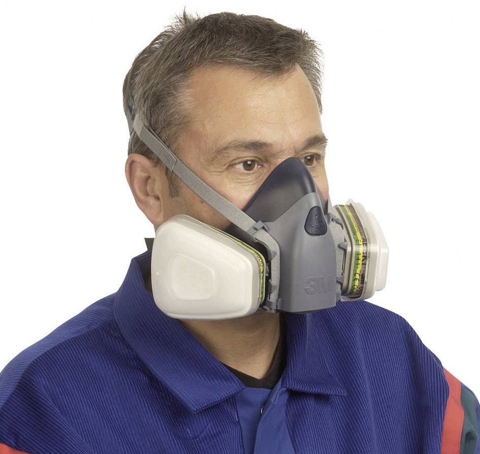En man bär en andningsskyddsmask med filterinsatser. Han har grått hår och är klädd i blå skyddsutrustning. Masken täcker mun och näsa.