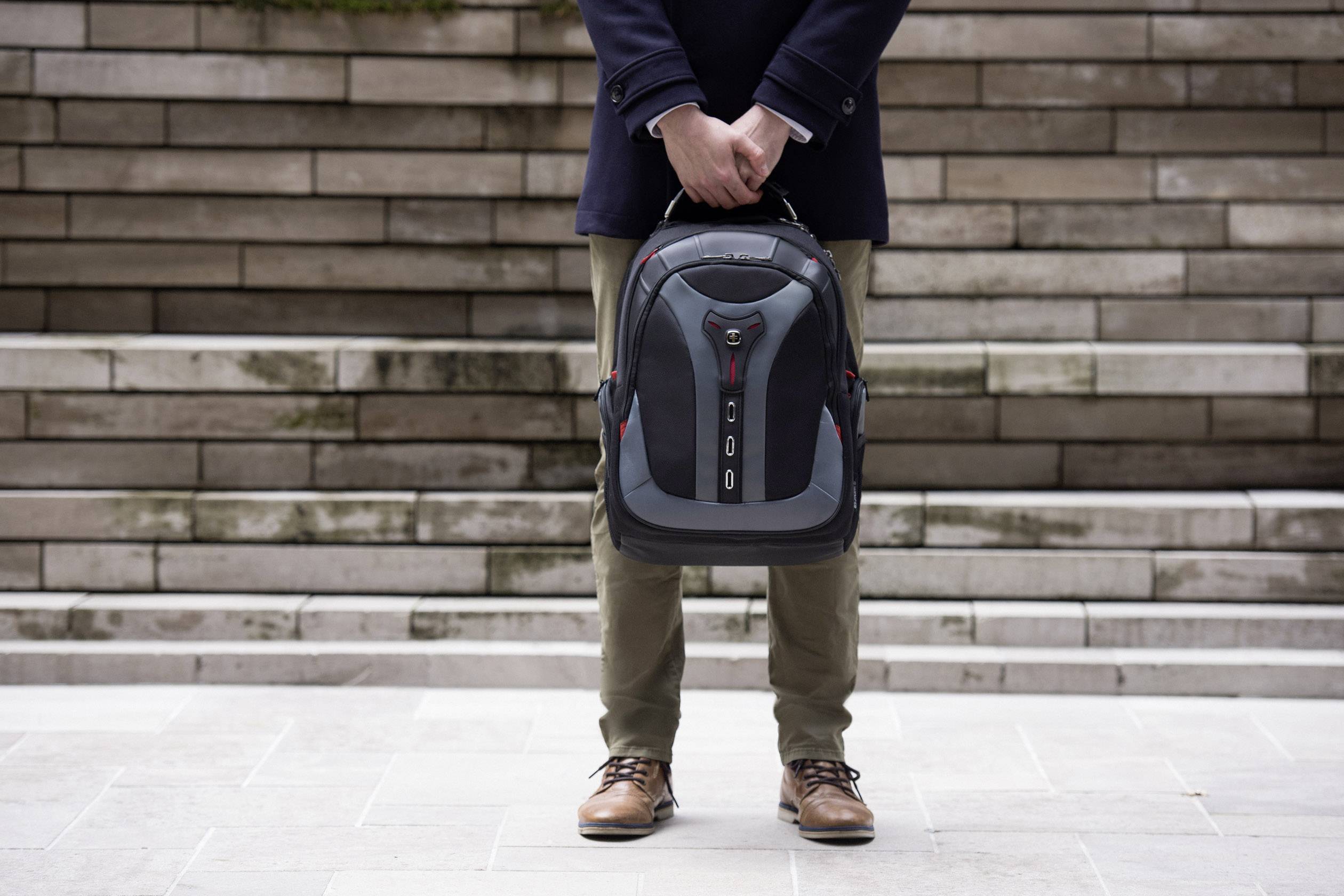 En person håller en ryggsäck framför sig. Bakgrund med stentrappor. Personen bär bruna skor och en mörk rock.