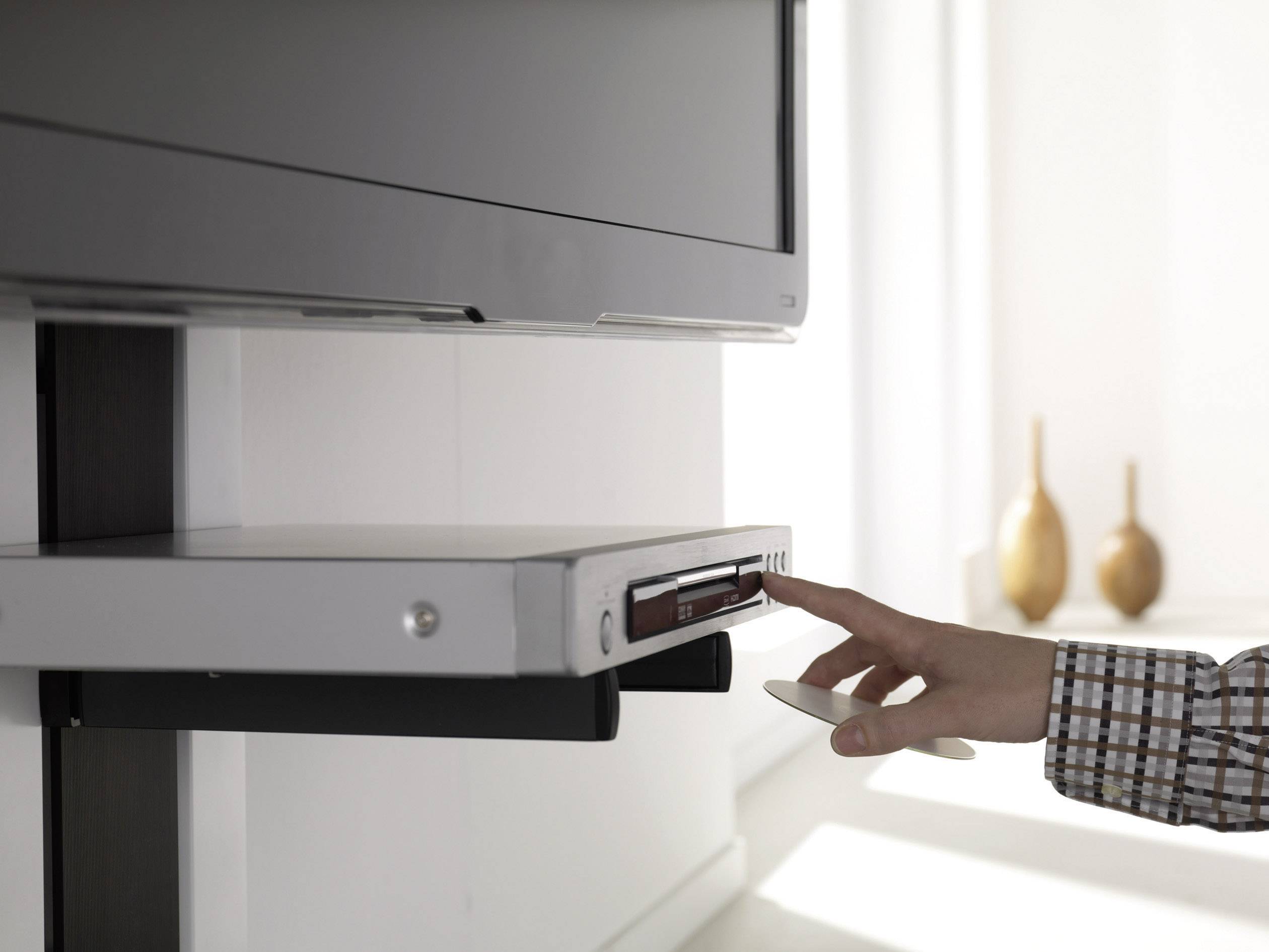 En hand manövrerar en DVD-spelare som är monterad under en väggmonterad TV. I bakgrunden syns två dekorativa vaser.