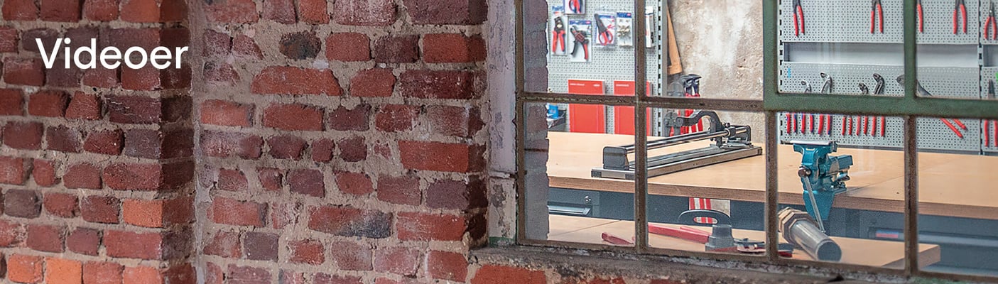 Et kig gennem et gammelt metalrammet vindue ind i et værksted med værktøj og en fliseskærer. Ordet "Videoer" står skrevet på murstensvæggen.