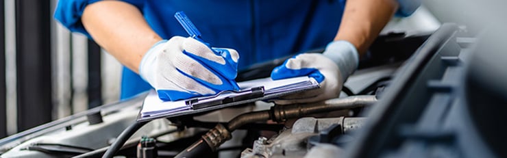 Automechaniker in blauem Overall und blauen Arbeitshandschuhen hält Stift und Clipboard, während er bei geöffneter Motorhaube den Motor eines Autos inspiziert und Notizen macht.