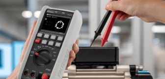 Eine Hand hält ein digitales Multimeter von Voltcraft, während eine andere Person mit einer roten und einer schwarzen Prüfspitze eine Messung an einem technischen Bauteil vornimmt.