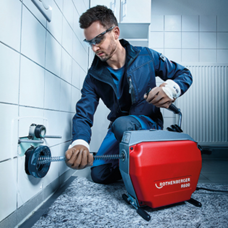 Ein Handwerker mit Schutzbrille führt die Reinigungsspirale einer elektrischen Rohrreinigungsmaschine in einen Abflussanschluss in einer gefliesten Wand ein.
