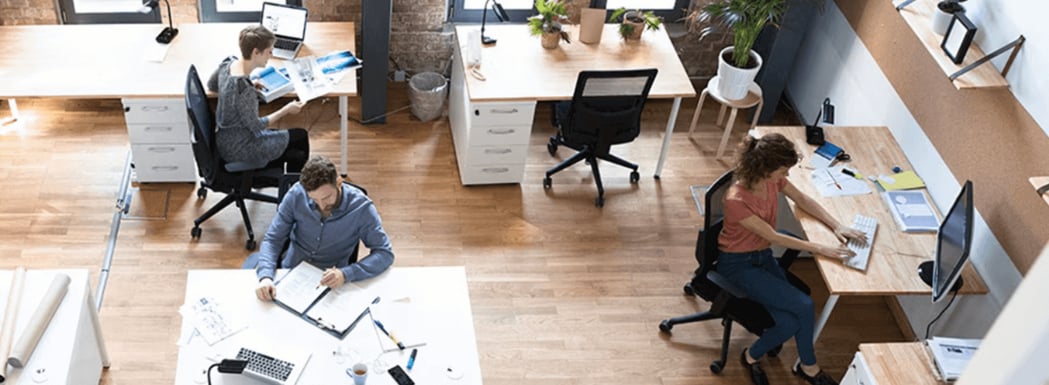 Eine Vogelperspektive zeigt ein modernes Büroraum mit vier Personen, die an verschiedenen Schreibtischen mit Computern und Unterlagen arbeiten.