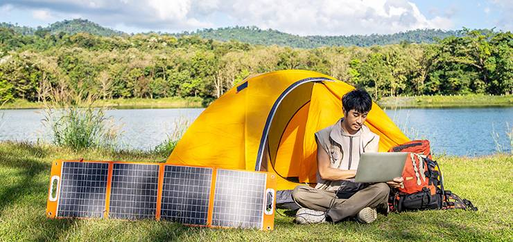 Person arbeitet am Laptop vor einem Zelt am See, mit mobilem Solarpanel für autarke Stromversorgung beim Camping.