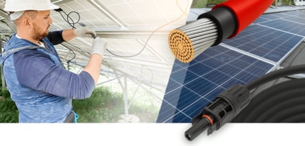  Ein Installateur in einem weißen Helm arbeitet an der Verkabelung von Solarpanelen, während im Vordergrund eine Nahaufnahme eines dicken, roten Stromkabels und eines schwarzen Steckers zu sehen ist.