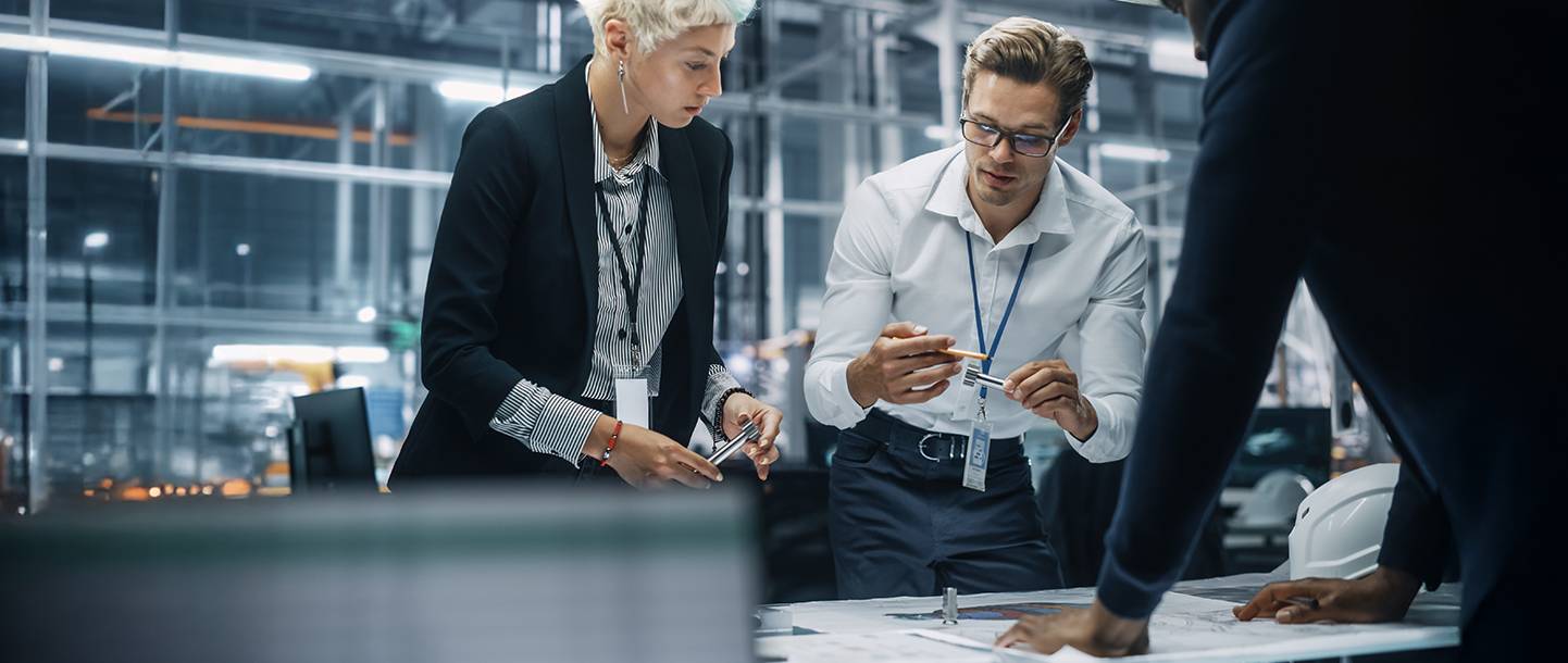 Personen besprechen ein Dokument auf dem Tisch, industrielle Umgebung im Hintergrund.