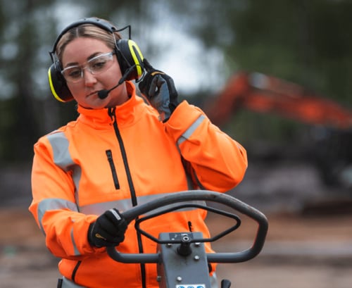 Eine Bauarbeiterin in Warnschutzjacke und mit Schutzbrille bedient eine Baumaschine, während sie einen Kapselgehörschützer mit Mikrofon am Kopf trägt und anfasst.