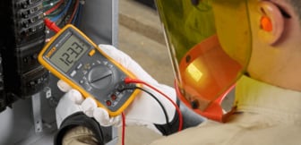 Ein Elektriker mit Schutzausrüstung hält ein gelbes Fluke-Multimeter, das eine Spannung von 123,3 V anzeigt, während er Messungen an einem Schaltschrank vornimmt.