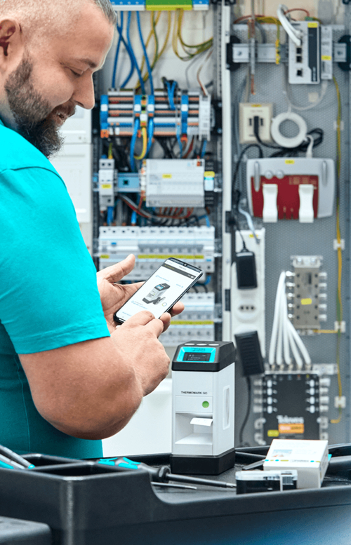 Ein Techniker steuert den Thermomark Go Beschriftungsdrucker mit seinem Smartphone vor einem Schaltschrank.