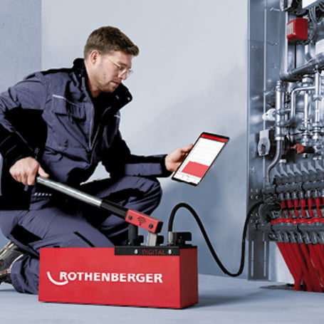 Ein Handwerker bedient eine rote digitale Prüfpumpe an einem Heizungsverteiler und kontrolliert die Messwerte auf einem Tablet.